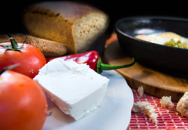 A simple breakfast with feta cheese in the foreground, tomatoes, peppers and bread, as well as a frying pan with fried eggs on a dark background.