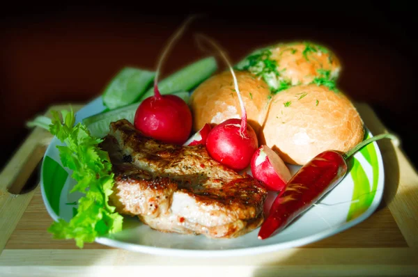 Gegrilltes Steak in einem Teller mit Paprika, Rettich, Gurke, Salat und Brot auf einem Holzbrett und dunklem Hintergrund. — Stockfoto