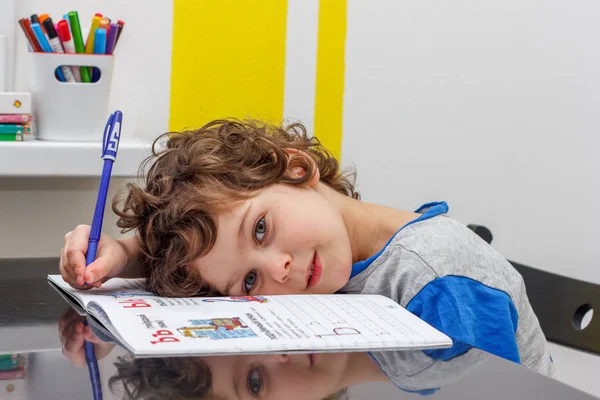 Уставший маленький мальчик, сидящий за столом и записывающий в блокнот во время домашней работы. Концепция большого количества заданий в школе — стоковое фото