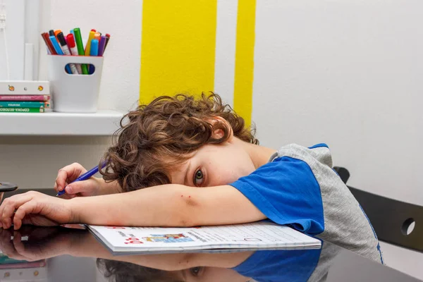 Уставший маленький мальчик сидел за столом, с головой на руке, записывая в блокнот, пока делал домашнее задание. Концепция большого количества заданий в школе — стоковое фото