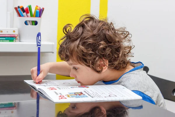 Уставший маленький мальчик, сидящий за столом и записывающий в блокнот во время домашней работы. Концепция большого количества заданий в школе — стоковое фото