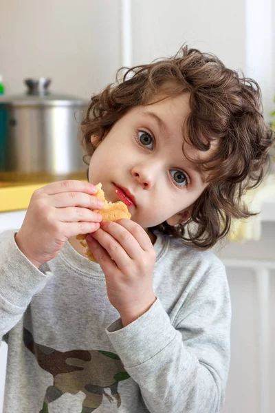 Un enfant frisé de cinq ans mange des biscuits avec appétit dans la cuisine — Photo