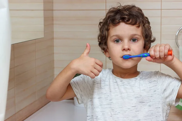 Lindo niño está cepillándose los dientes con cepillo de dientes y mostrando el pulgar hacia arriba. El concepto de salud infantil, medicina . —  Fotos de Stock