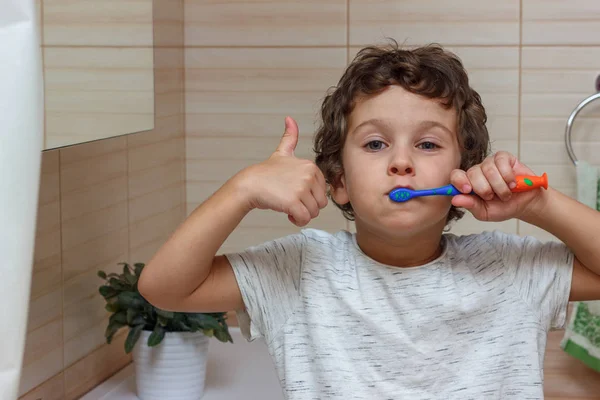 Lindo niño está cepillándose los dientes con cepillo de dientes y mostrando el pulgar hacia arriba. El concepto de salud infantil, medicina . —  Fotos de Stock
