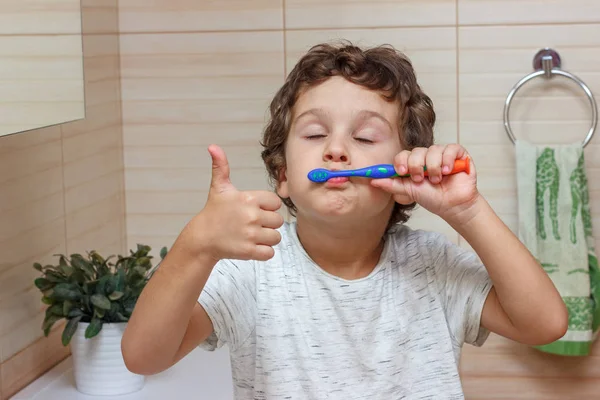 Lindo niño está cepillándose los dientes con cepillo de dientes y mostrando el pulgar hacia arriba. El concepto de salud infantil, medicina . —  Fotos de Stock