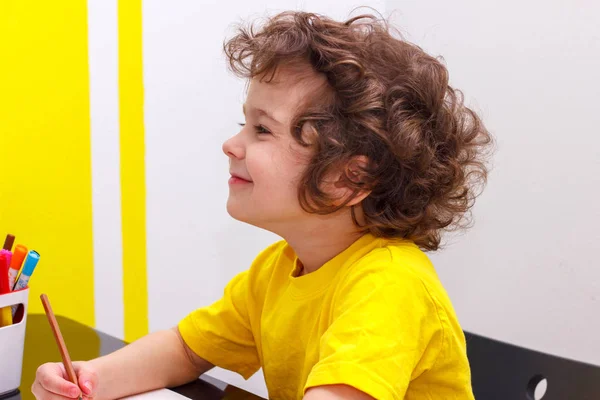 Маленький кудрявый мальчик в жёлтой футболке сидит за столом, улыбается, отдыхает после письма, глаза закрыты . — стоковое фото