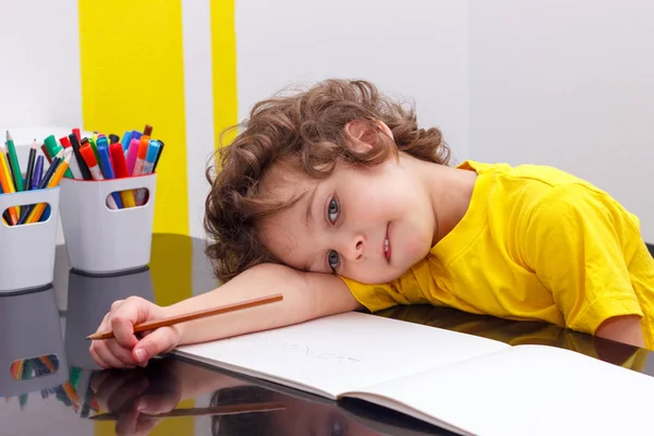 Уставший маленький мальчик сидит за столом, с головой на руке, пишет в блокноте, делая домашнее задание . — стоковое фото