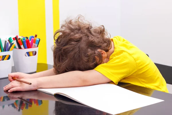 Уставший маленький мальчик сидит за столом, с головой на руке, пишет в блокноте, делая домашнее задание — стоковое фото