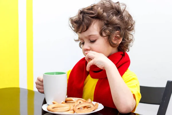 Petit garçon bouclé buvant dans une tasse de thé chaud par temps froid, assis à la maison dans l'écharpe dans la cuisine à la table — Photo