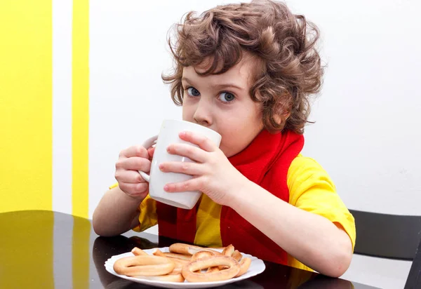 Petit garçon bouclé buvant dans une tasse de thé chaud par temps froid, assis à la maison dans l'écharpe dans la cuisine à la table — Photo