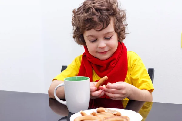 Petit garçon bouclé buvant dans une tasse de thé chaud par temps froid, assis à la maison dans l'écharpe dans la cuisine à la table — Photo