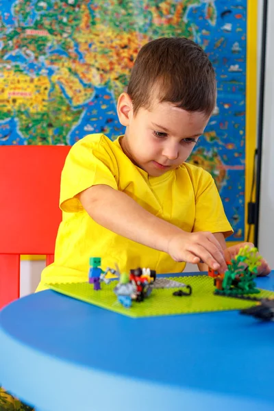 Маленький мальчик в жёлтой футболке играет с кучей разноцветных пластиковых блоков в помещении. Ребенок веселится со строительством и созданием — стоковое фото