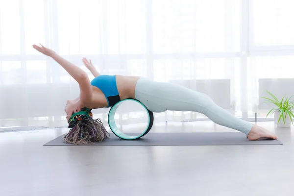 Yoga Indoor. Sports recreation. Beautiful young woman with dreadlocks in asana with yoga ring in room with big panoramic window. Wheel ring for yoga helps relieve pain in spine