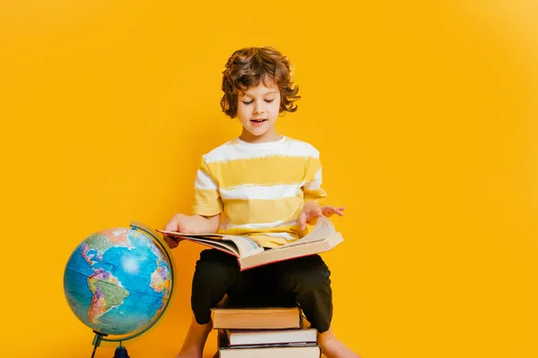 Кудрявый мальчик в очках и полосах футболке и сидит на стопке книг, рядом с шаром на желтом фоне пространства для текста. — стоковое фото