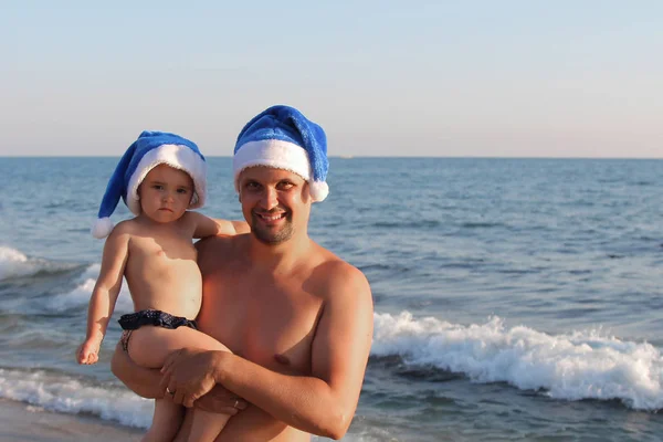 一个人抱着一个抱着一个戴着蓝色圣诞帽的小孩 戴着海洋的背景 — 图库照片