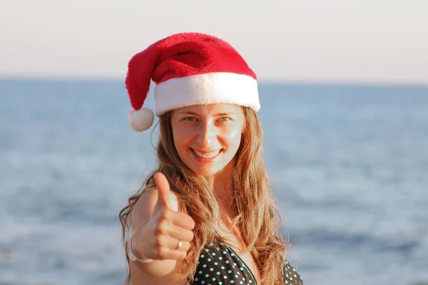 크리스마스 모자에 바다의 배경에 만족의 표시로 그녀의 엄지를 보여줍니다 — 스톡 사진