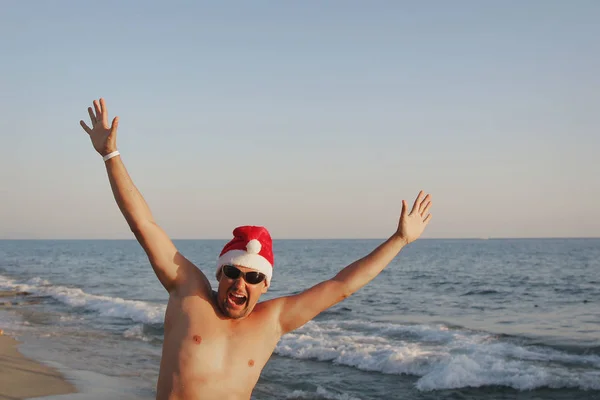 一个戴着红色圣诞帽 戴着太阳镜 双手举在海面上的人 — 图库照片