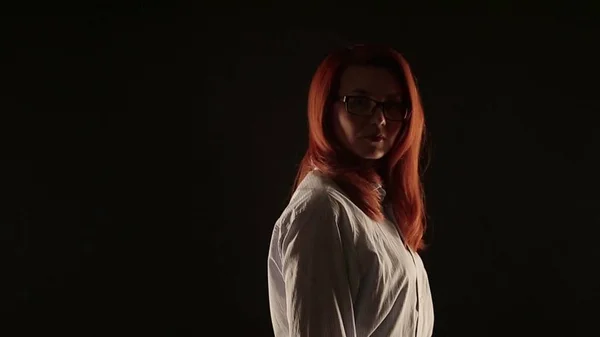 Uma menina bonita com cabelo vermelho em uma camisa branca posando em um fundo preto na frente da câmera. o modelo anuncia óculos, lentes ou serviços para restaurar a visão — Fotografia de Stock