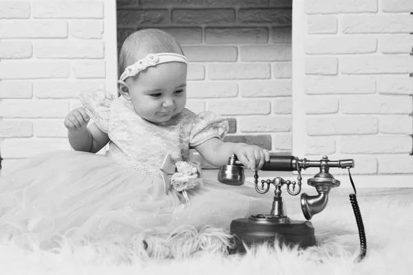 A little girl in a white dress sitting by the fireplace next to an old phone — Stock Photo, Image