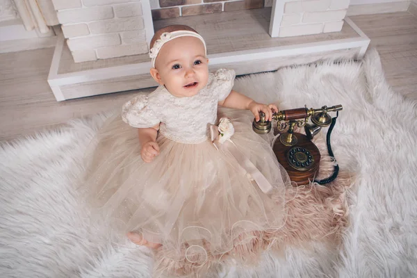 A little girl in a white dress sitting by the fireplace next to an old phone — Stock Photo, Image