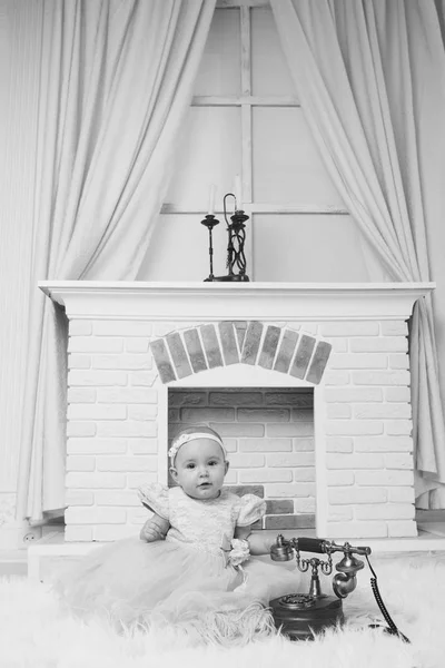 Una niña en un vestido blanco sentado junto a la chimenea junto a un teléfono viejo —  Fotos de Stock