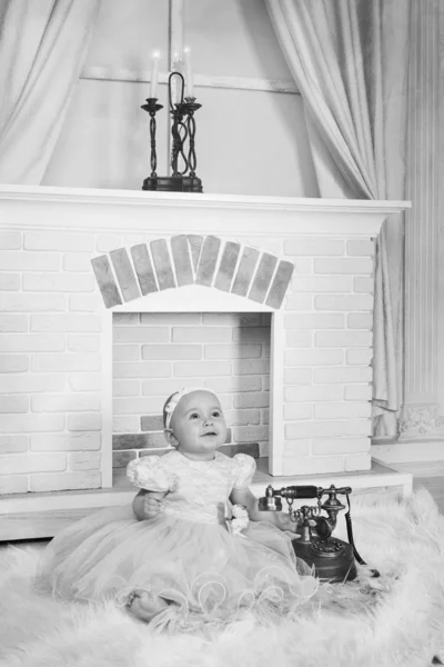 Ένα μικρό κορίτσι σε ένα λευκό φόρεμα κάθεται δίπλα στο τζάκι δίπλα σε ένα παλιό τηλέφωνο — Φωτογραφία Αρχείου