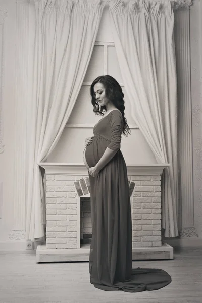 Uzun bir elbise güzel hamile kız, ev iç arka plan karşı karnına dokunmadan: şömine ve perdeler — Stok fotoğraf