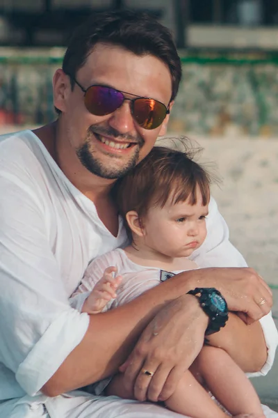 Papa en lunettes de soleil tient le bébé dans ses bras sur le fond de la plage. Le concept de l'éducation du père de jeunes enfants, Bonne enfance, une famille amicale . — Photo