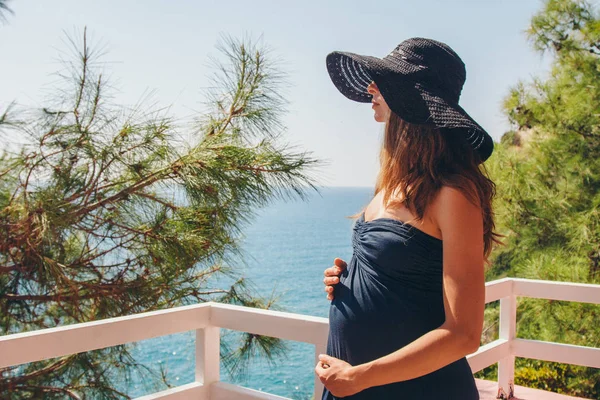 Begreppet turism, fritid är en vacker gravid flicka med långt hår, sola i den svarta klänningen och hatt på balkongen på hotellet med utsikt över tropiska växter och en liten byggnad — Stockfoto