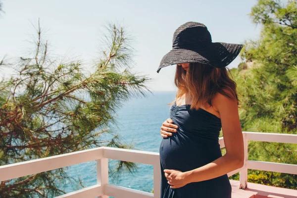 El concepto de turismo, el ocio es una hermosa chica embarazada con el pelo largo, tomando el sol en el vestido negro y sombrero en el balcón del hotel con vistas a las plantas tropicales y un pequeño edificio — Foto de Stock