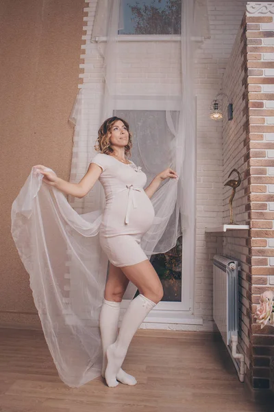 Uma mulher grávida em um vestido leve e meias fica pensativo na janela, brincando com cortinas. O conceito de um estilo de vida saudável, IVF — Fotografia de Stock