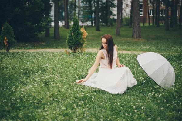 Una bella bruna in un vestito bianco con un ombrello si siede sull'erba. Bella ragazza in natura — Foto Stock