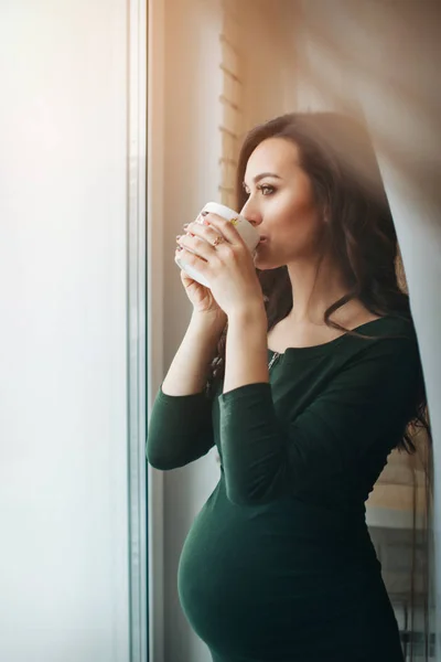 Portret kobiety ciężarnej z filiżanką w ręku w oknie. Spokojna przyszła matka. Przerwa na lunch lub kawę, herbatę — Zdjęcie stockowe