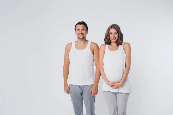 Gravid kvinna och man i sportkläder på vit bakgrund. Flickan håller magen med munnen vidöppen och ett leende på läpparna — Stockfoto