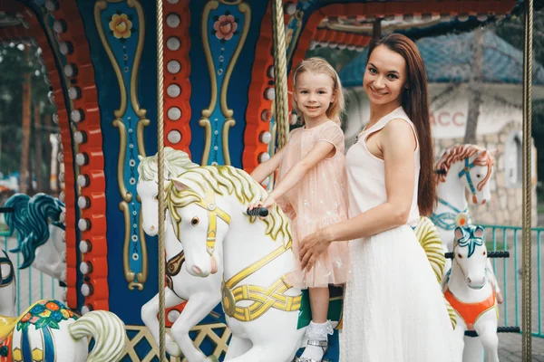 A little girl with her mother riding in the Park on a toy horse on the carousel. Entertainment industry concept, family day, children's parks, playgrounds — Stock Photo, Image