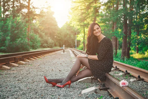 Una bella bruna in abito nero e scarpe rosse siede sulle rotaie. Donna pubblicizza accessori, cosmetici, trucco, vestiti — Foto Stock