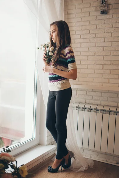 A beautiful girl without makeup with flowers looks out from behind the curtain, a woman at the window. The concept of advertising natural makeup, morning pastime