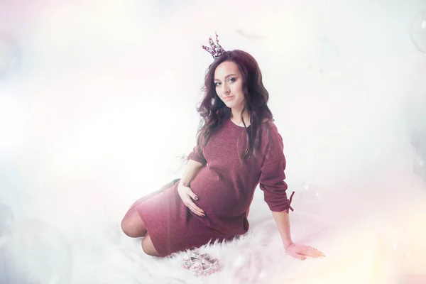 Hamile bir kadın karnında bebek paçası taşıyor. Taç içindeki prenses. Yeni doğanlar için moda. Çocuk ayakkabısı reklamı — Stok fotoğraf