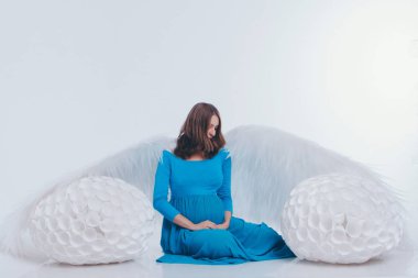 Mavi elbiseli, melek kanatlı hamile bir kız beyaz arka planda oturuyor. Bir melek kadının büyük beyaz topları vardır. Noel konsepti, mistik yaratıklar, Cadılar Bayramı