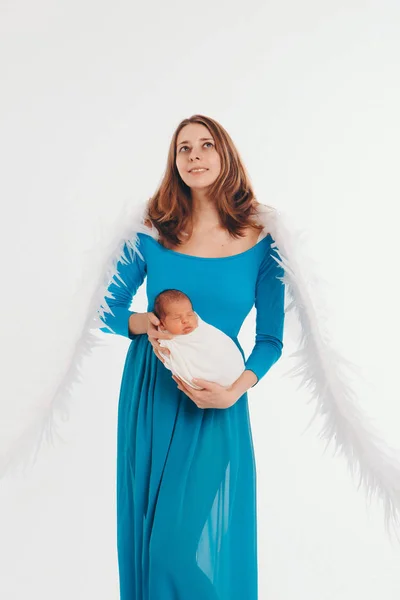 Egy lány kék ruhában, angyalszárnyakkal, kezében egy újszülöttel, fehér háttérrel. Egy angyali nőnek és egy gyereknek nagy fehér golyói vannak. A koncepció a karácsony, misztikus lények, Halloween — Stock Fotó
