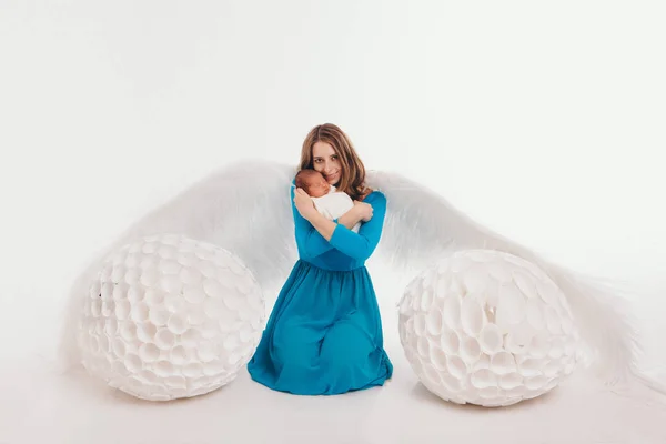 Девушка в синем платье с ангельскими крыльями, держащая новорожденного ребенка на белом фоне. У женщины-ангела и ребенка большие белые яйца. Концепция Рождества, мистические существа, Хэллоуин — стоковое фото