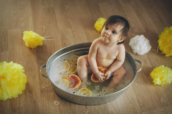 Een klein meisje baadt in een bad met citroen en grapefruit. Een kind wast zich in een bassin tussen gele bloemen. concept van kindertijd, gezondheidszorg, Ivf, hygiëne, citrustherapie — Stockfoto