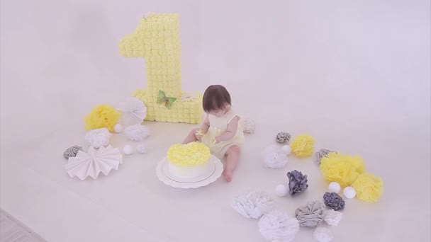 誕生日のお祝い 白い背景に彼女の手でケーキを食べる女の子 子供は食べ物に覆われています 甘酸っぱい 装飾の間の床に 数字1 人工の花と白いボール コピースペース 料理の概念 — ストック動画