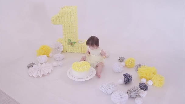 誕生日のお祝い 白い背景に彼女の手でケーキを食べる女の子 子供は食べ物に覆われています 甘酸っぱい 装飾の間の床に 数字1 人工の花と白いボール コピースペース 料理の概念 — ストック動画