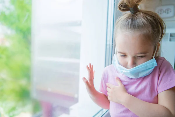 the girl in the medical mask leaned against the window. the girl looked out the window. a person is bored in self-isolation. I look at the world through glass. sad face