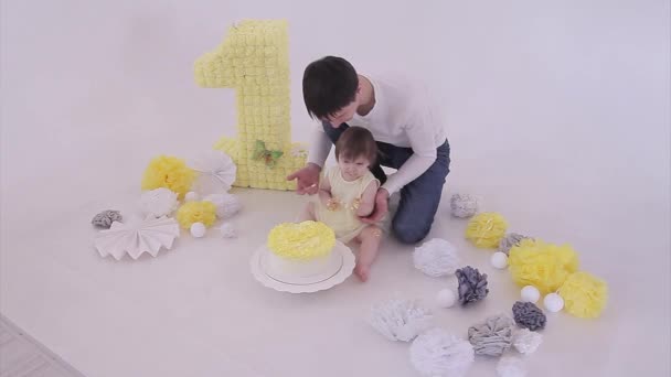 誕生日のお祝い 白い背景に彼女の手でケーキを食べる男と女の子 子供は食べ物に覆われています 甘酸っぱい 装飾の間の床に 数字1 人工の花と白いボール 誕生日 料理の概念 — ストック動画