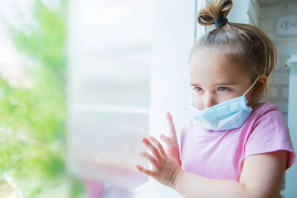 the girl in the medical mask leaned against the window. the girl looked out the window. a person is bored in self-isolation. I look at the world through glass. sad face