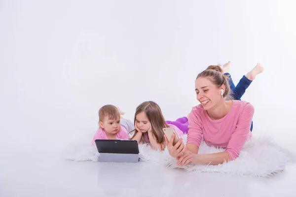 modern technologies in everyday life: a woman talks on the phone through a headset, children watch a cartoon on a tablet. Hobbies and recreation with gadgets. Family vacation, joint pastime. Parents with girls on the floor