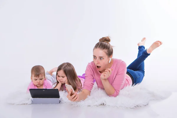 Modern technologies in everyday life: woman talks on phone through headset, children watch cartoon on tablet. Hobbies and recreation with gadgets. Family vacation. Parents with girls on floor