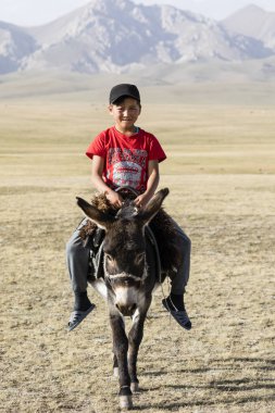 Şarkı Kul, Kırgızistan, 8 Ağustos 2018: Kırmızı bir tişört bir çocuk şarkı Kul Gölü Kırgızistan'daki bozkır aracılığıyla bir eşek rides